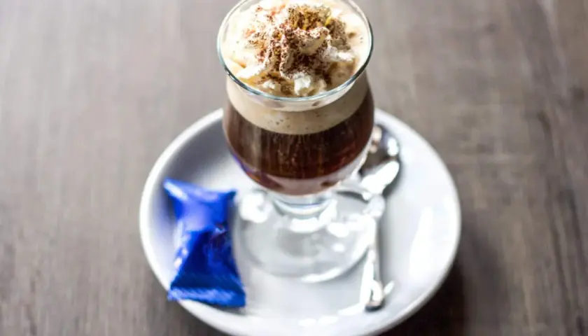 Ein Glas Kaffee mit Rum, gekrönt mit Sahne und Kakaopulver, serviert mit einer blauen Praline und Löffel auf einem Unterteller.