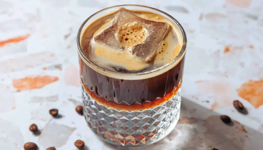 Erfrischender Espresso Tonic mit großem Eiswürfel und cremigem Schaum in einem Glas mit Rautenmuster. Kaffeebohnen liegen verstreut.