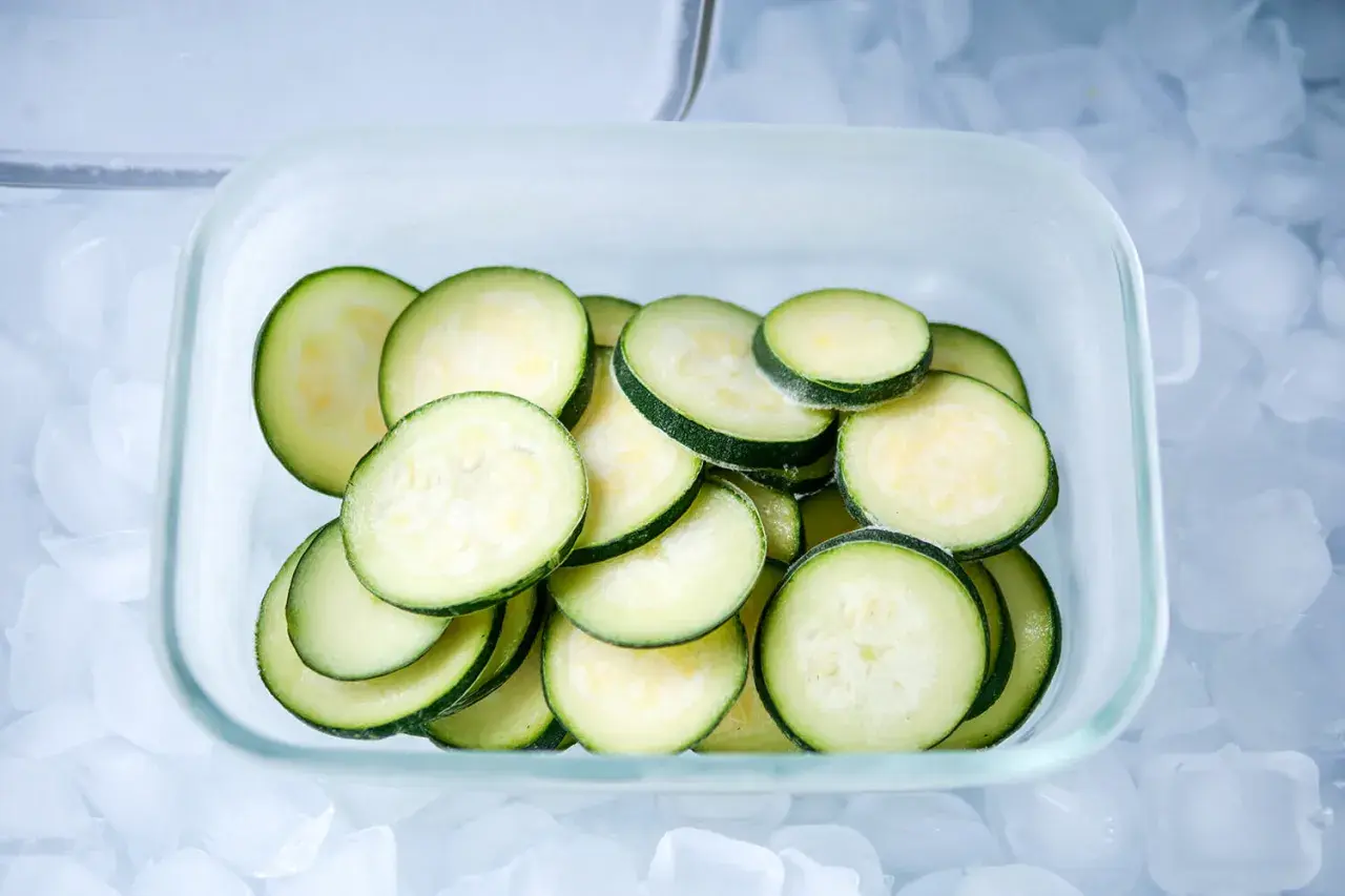 Scheiben von Zucchini lagern in einem Glasbehälter auf Eiswürfeln.