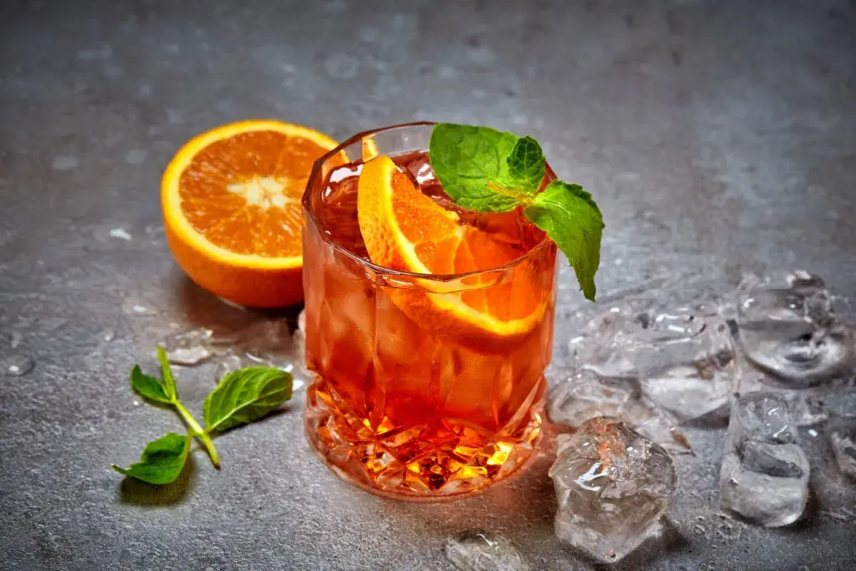 Ein erfrischender Cynar Spritz mit Orangenscheiben und Minze, umgeben von Eiswürfeln auf grauem Untergrund.