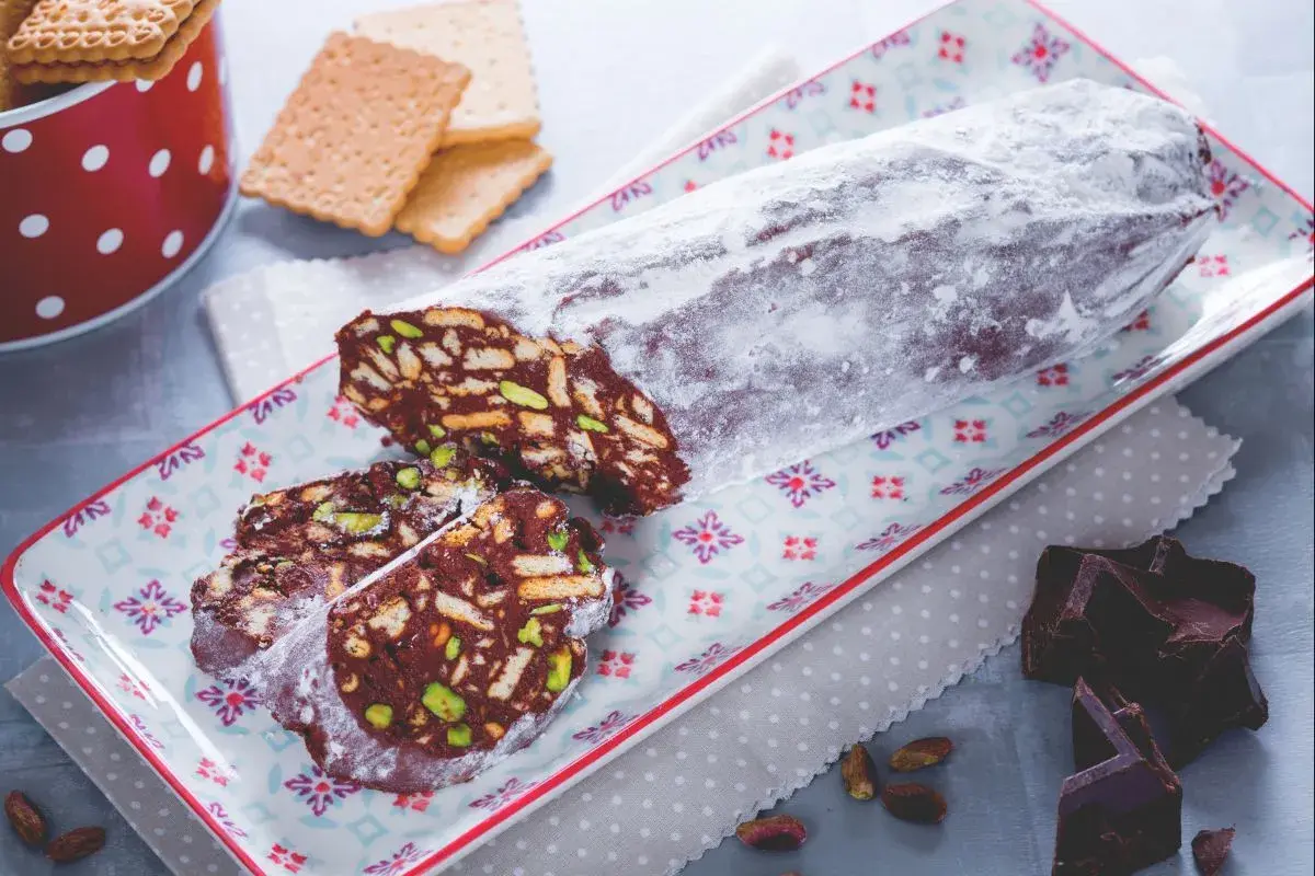 Ein köstlicher salame al cioccolato, bestäubt mit Puderzucker, auf einem bunten Teller. Daneben liegen zerbrochene Schokoladenstücke und Pistazien.