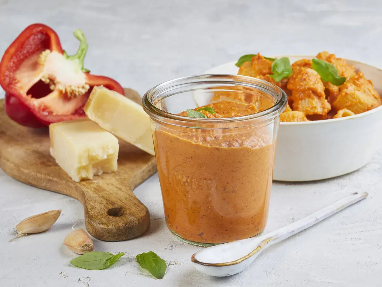 Ein Glas mit leuchtend rotem Paprika-Pesto steht neben einer Schüssel Pasta. Auf einem Holzbrett liegen eine halbe rote Paprika, Parmesankäse, Knoblauch und Basilikum.