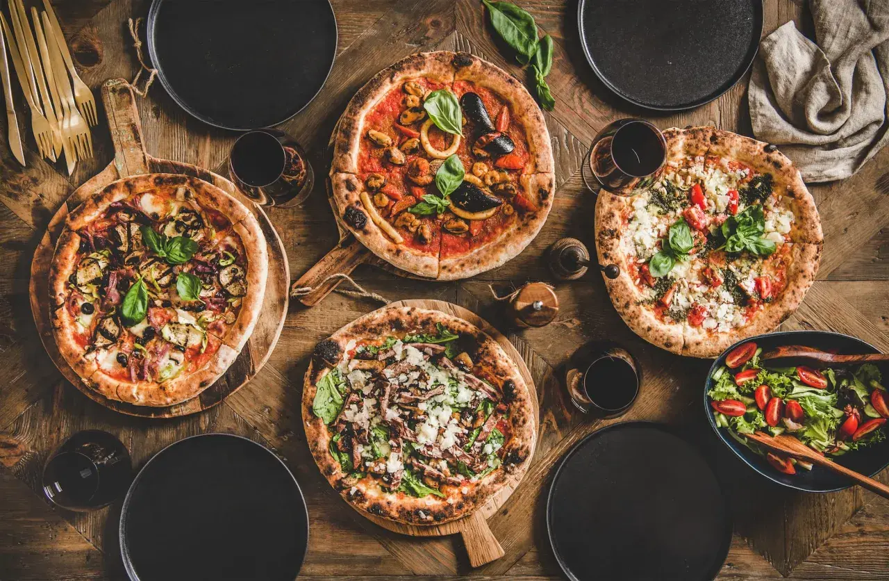 Vier leckere Pizzen, ein Salat und Rotwein auf rustikalem Holztisch. Perfekt für einen gemütlichen Abend mit Freunden.