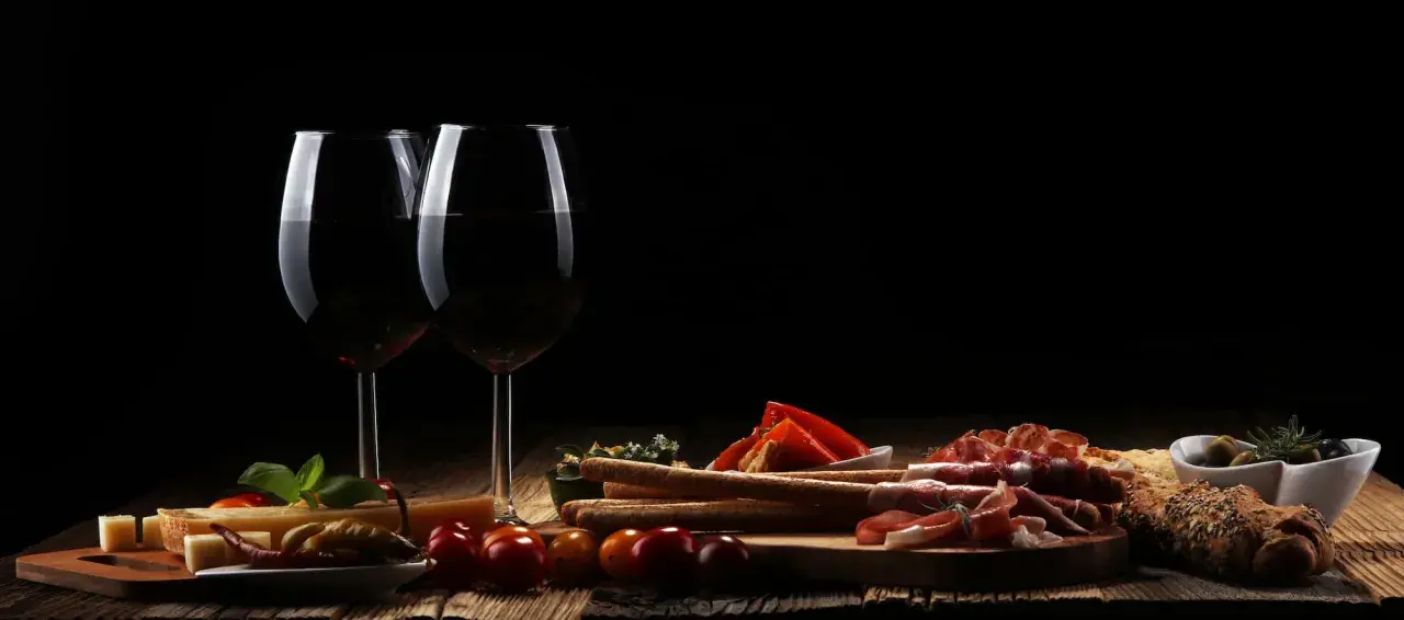 Zwei Gläser Rotwein, ein Cuvee Wein, stehen neben einer rustikalen Platte mit Käse, Schinken, Brot und Oliven.
