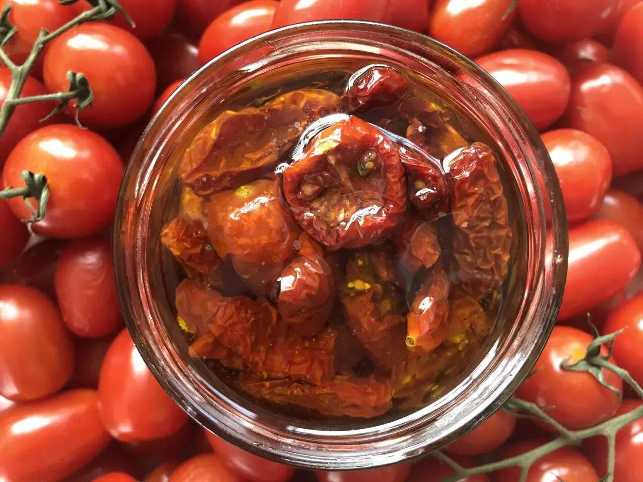 Ein Glas mit halbgetrockneten Tomaten, umgeben von frischen, roten Tomaten.