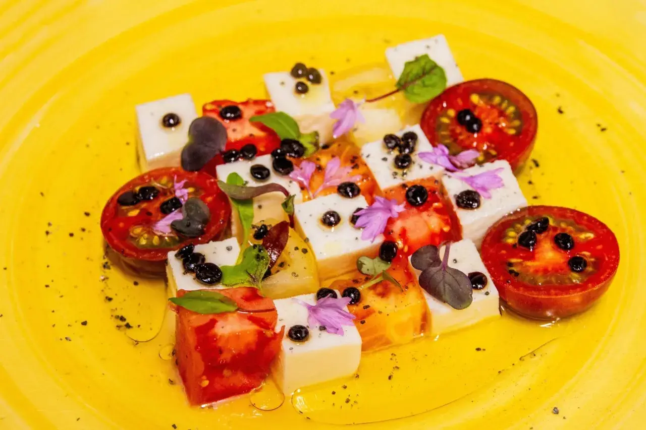 Tomaten-Mozzarella-Salat mit Balsamico-Kaviar, garniert mit Kräutern und Blüten auf gelbem Teller.