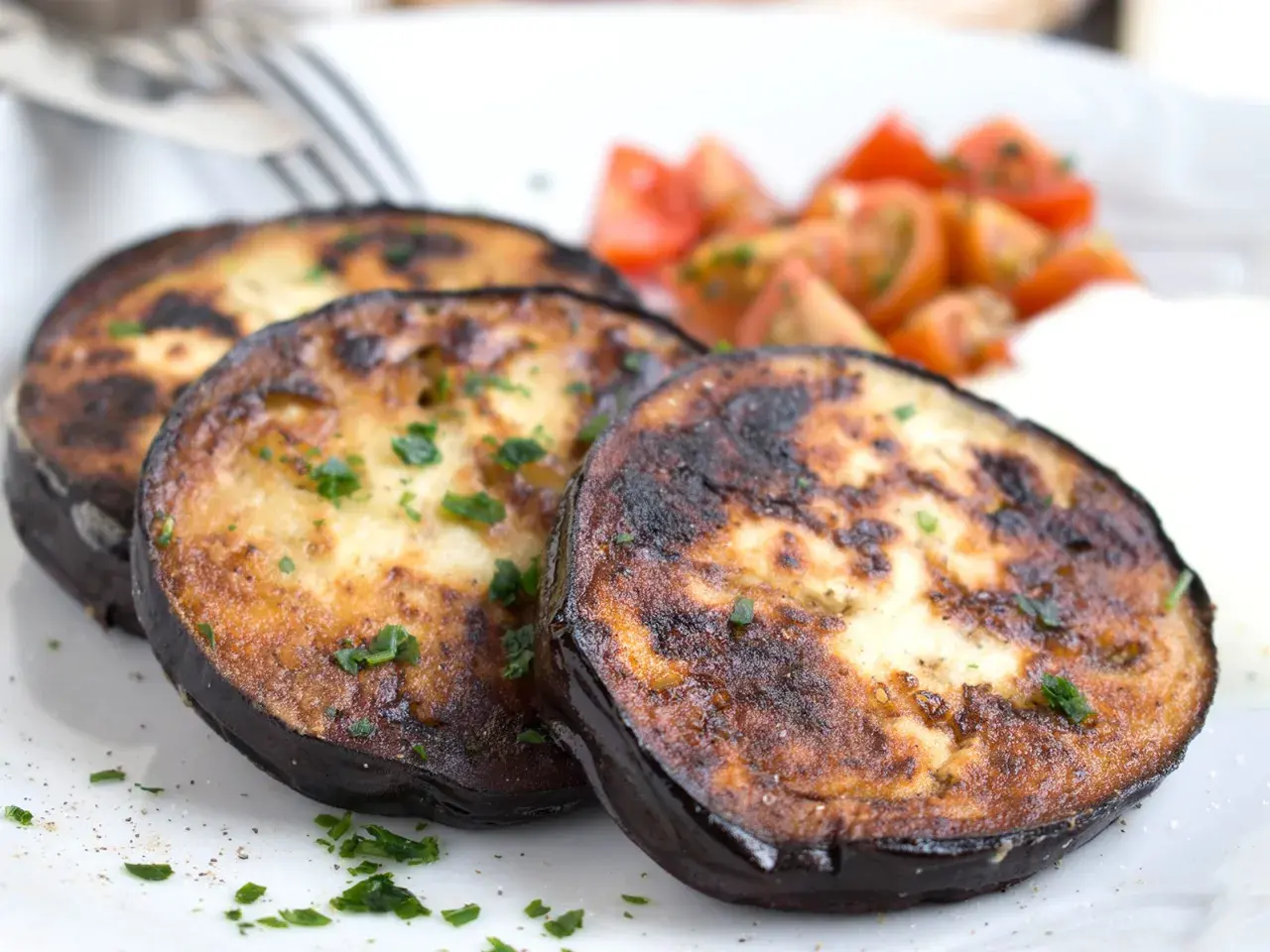 Drei Scheiben perfekt gebratene Aubergine, garniert mit Petersilie, serviert mit Kirschtomaten und einer cremigen Beilage.