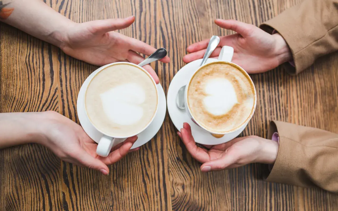 Zwei Hände reichen sich zwei Tassen mit cremigem Kaffee mit Milch, Latte Art in Herzform.