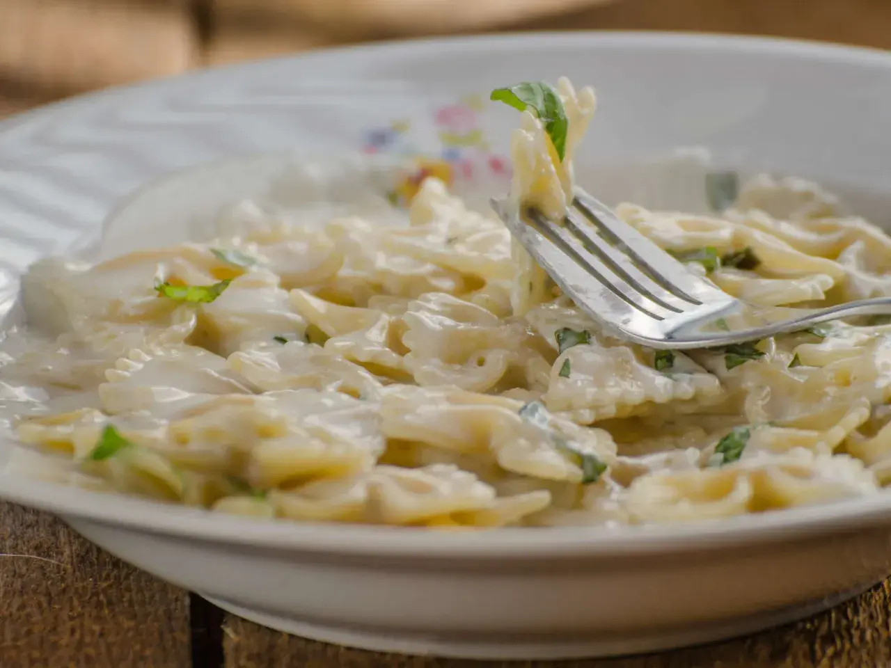 Farfalle-Nudeln in cremiger Sauce mit Zucchini und Ricotta, garniert mit frischen Kräutern. Ein Gabel hebt eine Portion an.