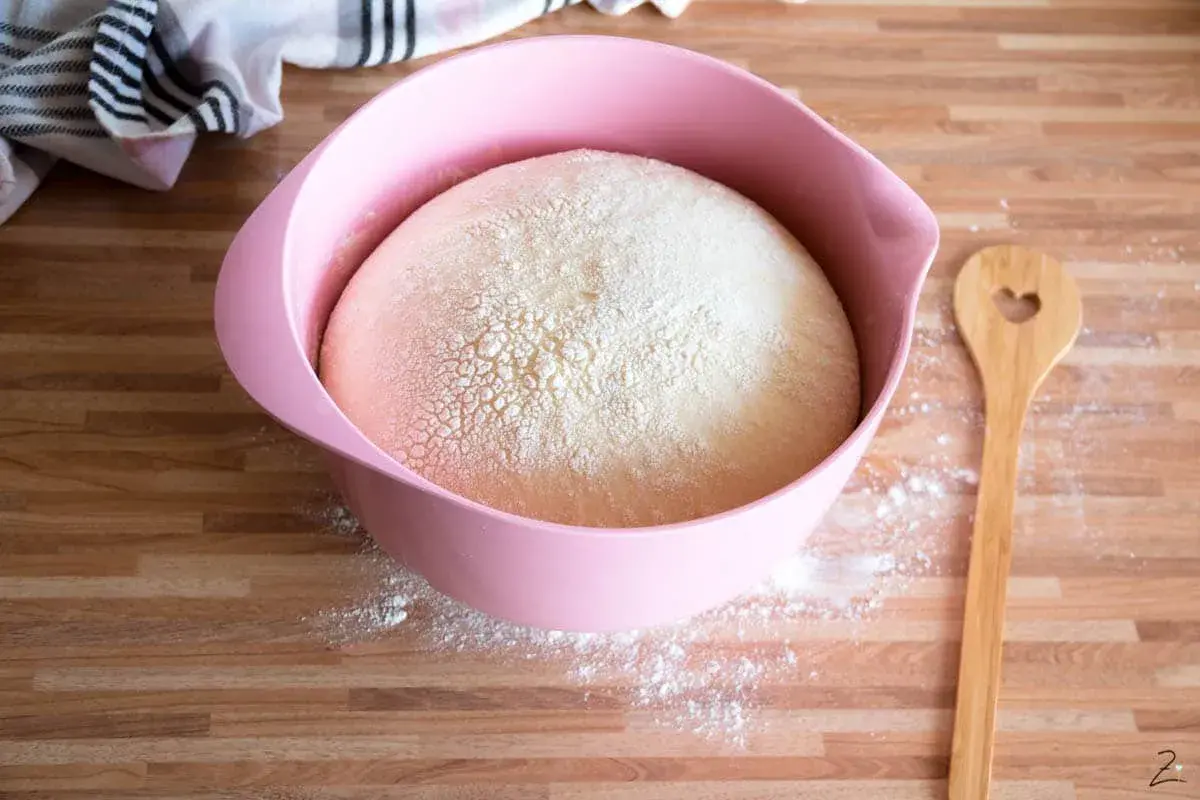 Ein aufgegangener Hefeteig in einer rosa Schüssel, bereit für die Zubereitung. Daneben liegt ein Holzlöffel mit Herz.