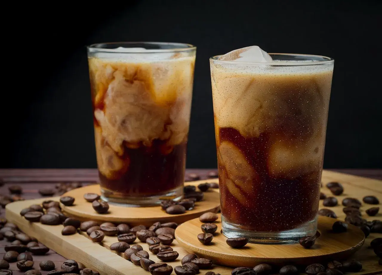Zwei Gläser mit cremigem, kaltem Kaffee, umgeben von gerösteten Kaffeebohnen. Ein erfrischender Genuss an einem heißen Tag.