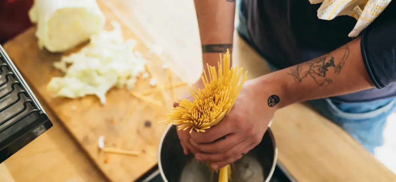Hände mit Tattoos halten Spaghetti über einen Topf, um Nudeln zu kochen. Auf dem Holztisch liegt geschnittener Kohl.