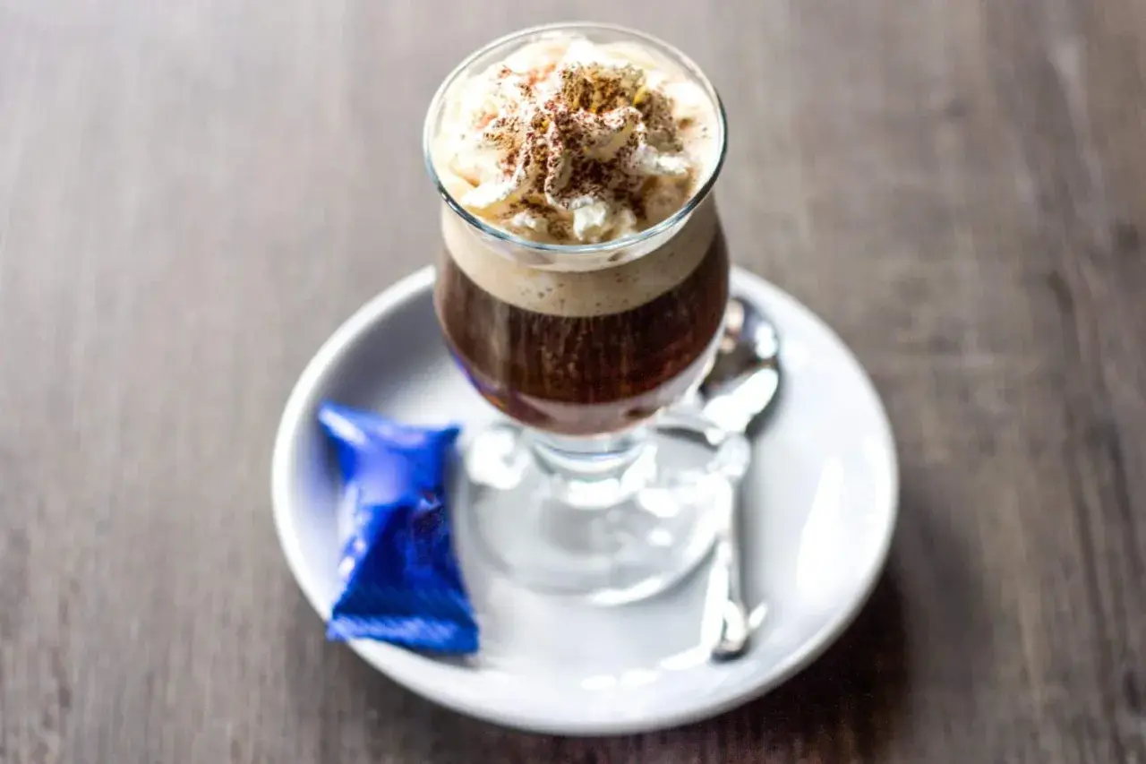Ein Glas Kaffee mit Rum, gekrönt mit Sahne und Kakaopulver, serviert mit einer blauen Praline und Löffel auf einem Unterteller.