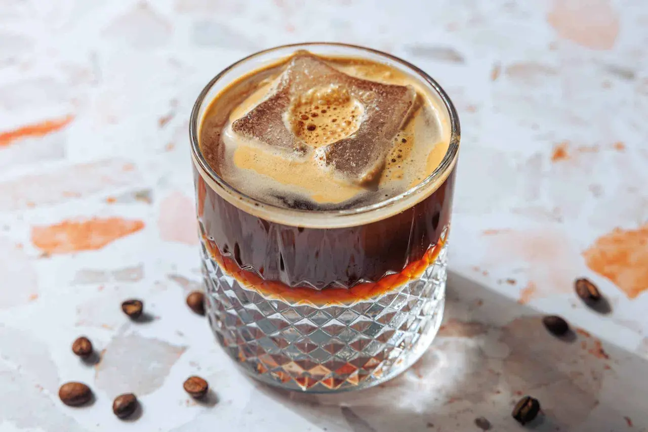 Erfrischender Espresso Tonic mit großem Eiswürfel und cremigem Schaum in einem Glas mit Rautenmuster. Kaffeebohnen liegen verstreut.