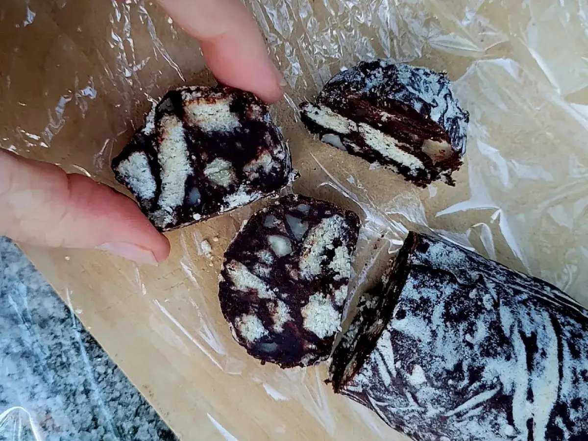 Schokoladensalami, ein köstlicher Nachtisch aus Keksen und Schokolade, wird von Fingern gehalten und in Scheiben geschnitten.