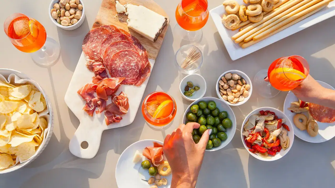 Ein Tisch voller K&ouml;stlichkeiten f&uuml;r einen italienischen Aperitif: Salami, Schinken, K&auml;se, Oliven, Chips, Grissini und erfrischende Aperol Spritz.