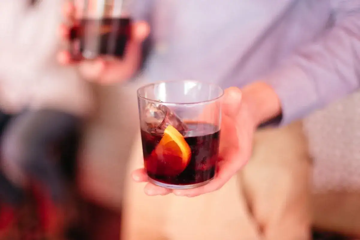 Eine Hand h&auml;lt ein Glas mit einem dunklen Getr&auml;nk, Eis und einer Orangenscheibe. So h&auml;lt man ein Weinglas richtig.