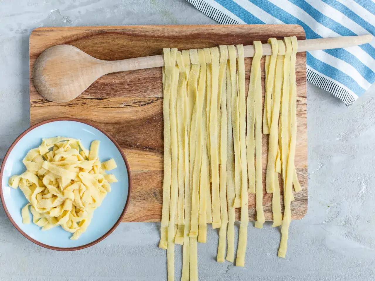 Frische Bandnudeln h&auml;ngen zum Trocknen &uuml;ber einem Holzl&ouml;ffel. Ein Teller mit bereits gekochten Nudeln liegt daneben. Perfekt f&uuml;r ein Bandnudeln Rezept.