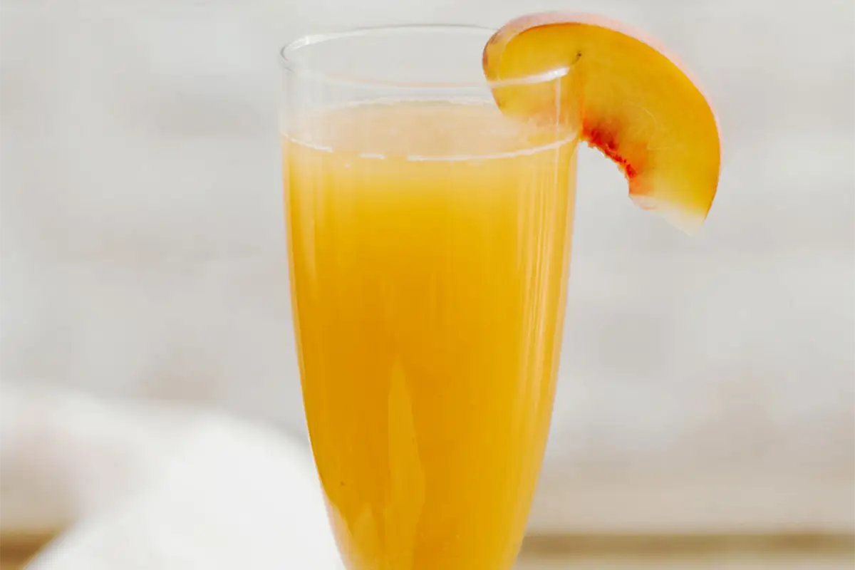 Ein Glas mit einem leuchtend orangen Getr&auml;nk, garniert mit einem Pfirsichspalt. Das ist das Bellini Rezept original.