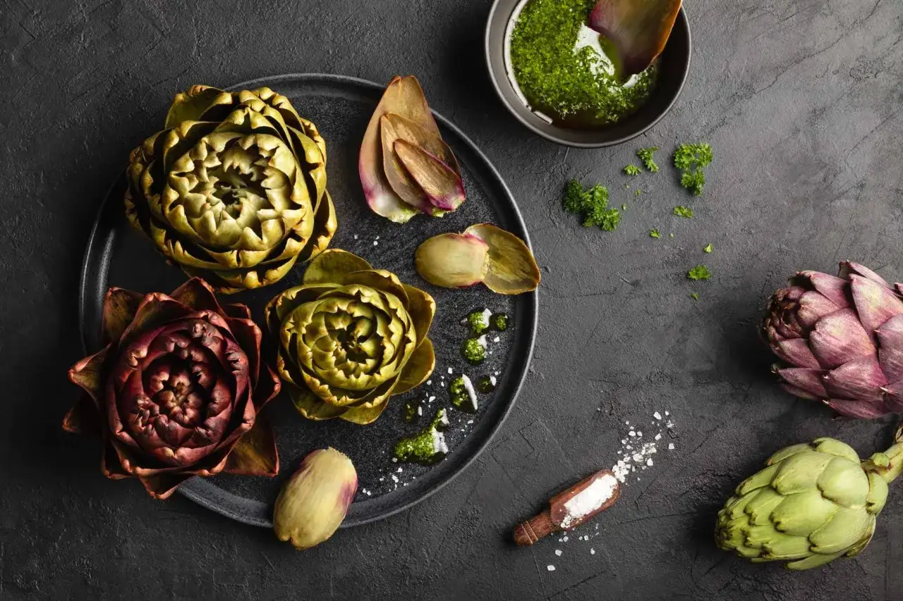 Frische Artischocken zum Kochen, mit Kr&auml;uter-Dip und grobem Salz auf dunklem Untergrund arrangiert.