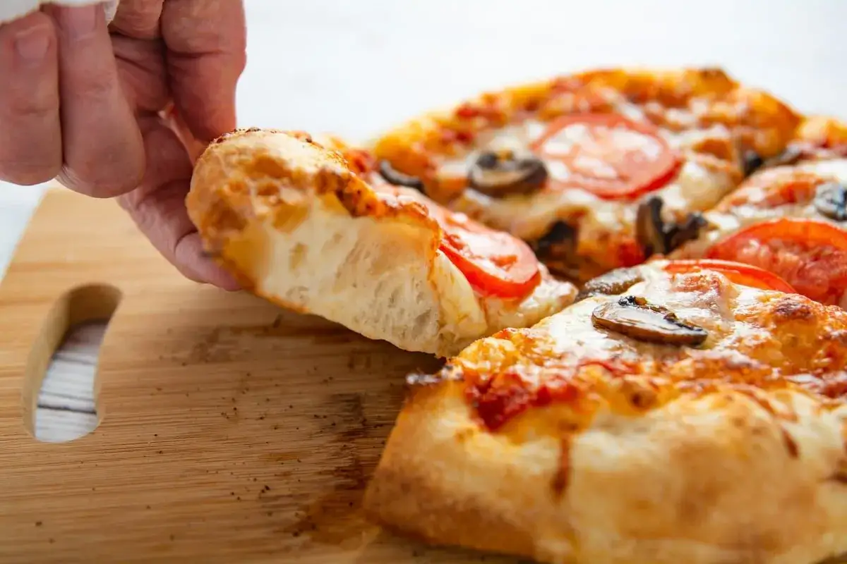 Ein St&uuml;ck Pizza mit Tomaten und Pilzen wird von einer Hand hochgehoben. Der fluffige Teig zeigt, wie man einen hefeteig macht.