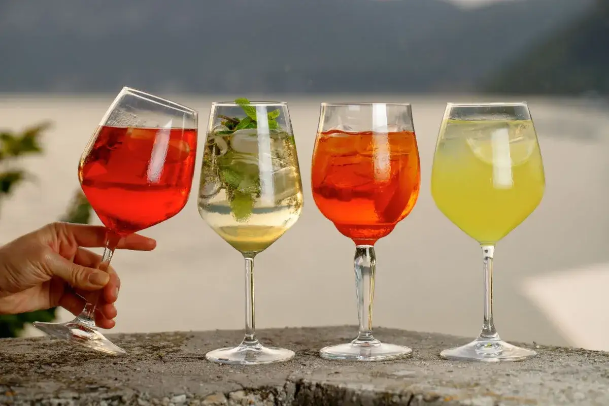 Vier bunte Aperitifs, darunter ein Cynar Spritz, stehen auf einer Steinmauer mit Blick auf das Wasser.