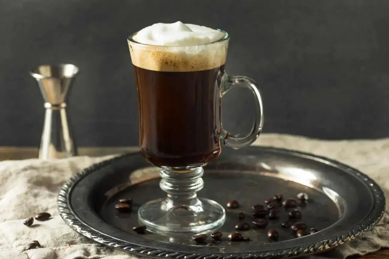 Ein Glas heißer Kaffee mit einer cremigen Haube, bereit für einen Schuss. Kaffeebohnen liegen auf einem Silbertablett.