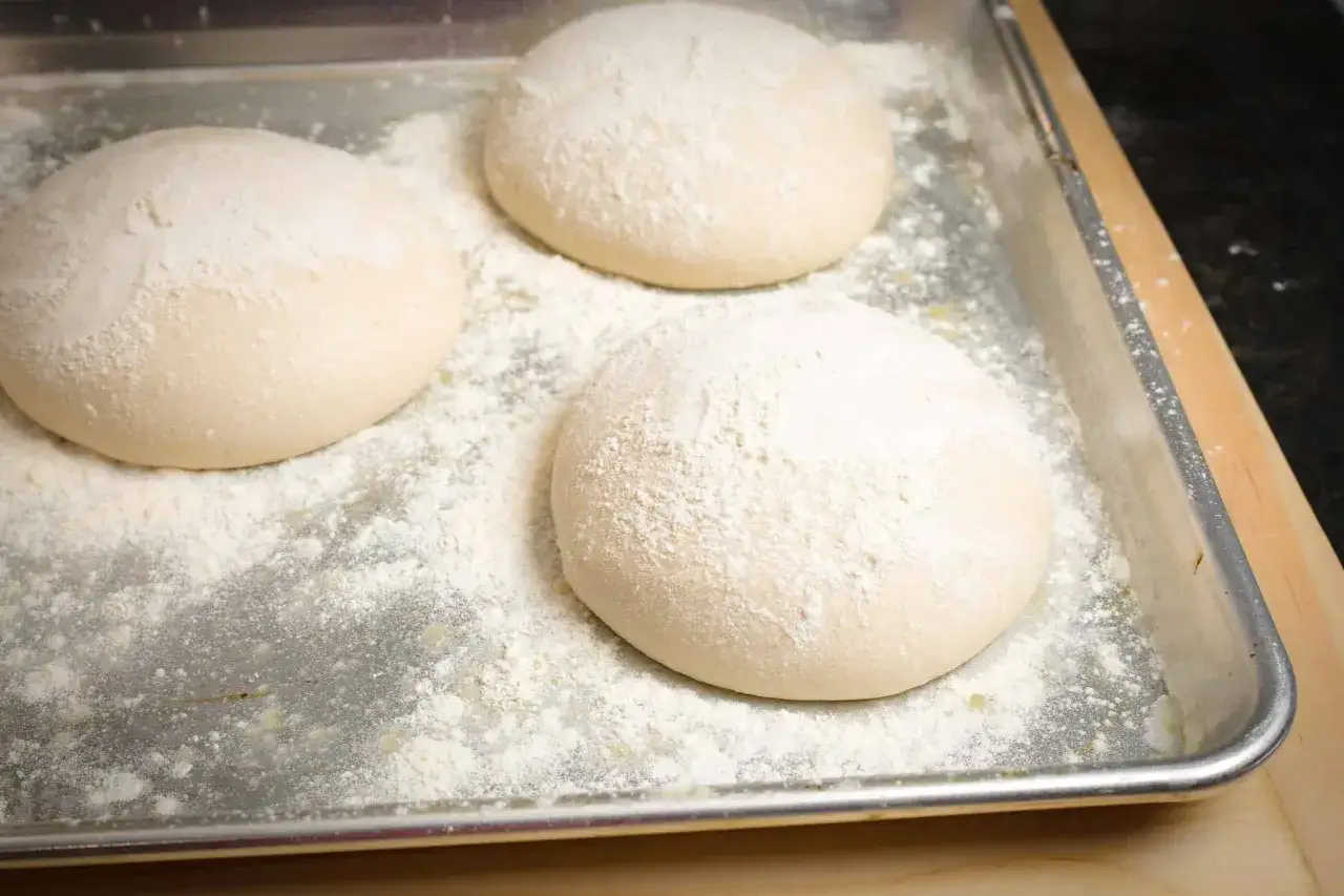 Drei runde Teiglinge für Pizzaböden liegen auf einem Backblech, bestäubt mit Mehl. Der Pizzateig Napoletana wartet auf seine Zubereitung.
