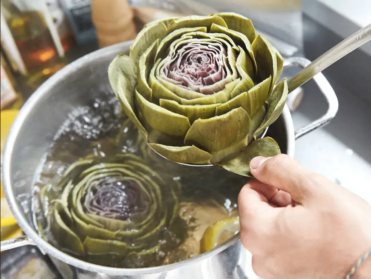 Artischocken zubereiten: Eine Hand hebt eine dampfende Artischocke mit einer Schöpfkelle aus kochendem Wasser. Eine weitere Artischocke kocht bereits im Topf.