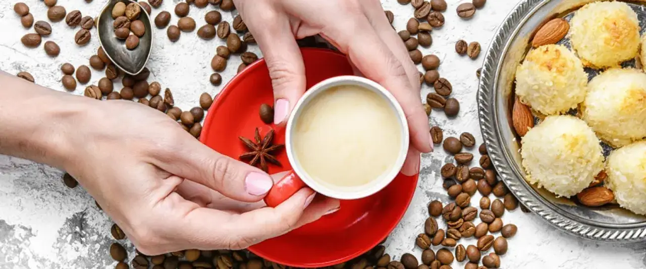Hände halten eine Tasse Kaffee, umgeben von Kaffeebohnen und Kokosmakronen. Inspiration für neue Kaffee Rezepte.