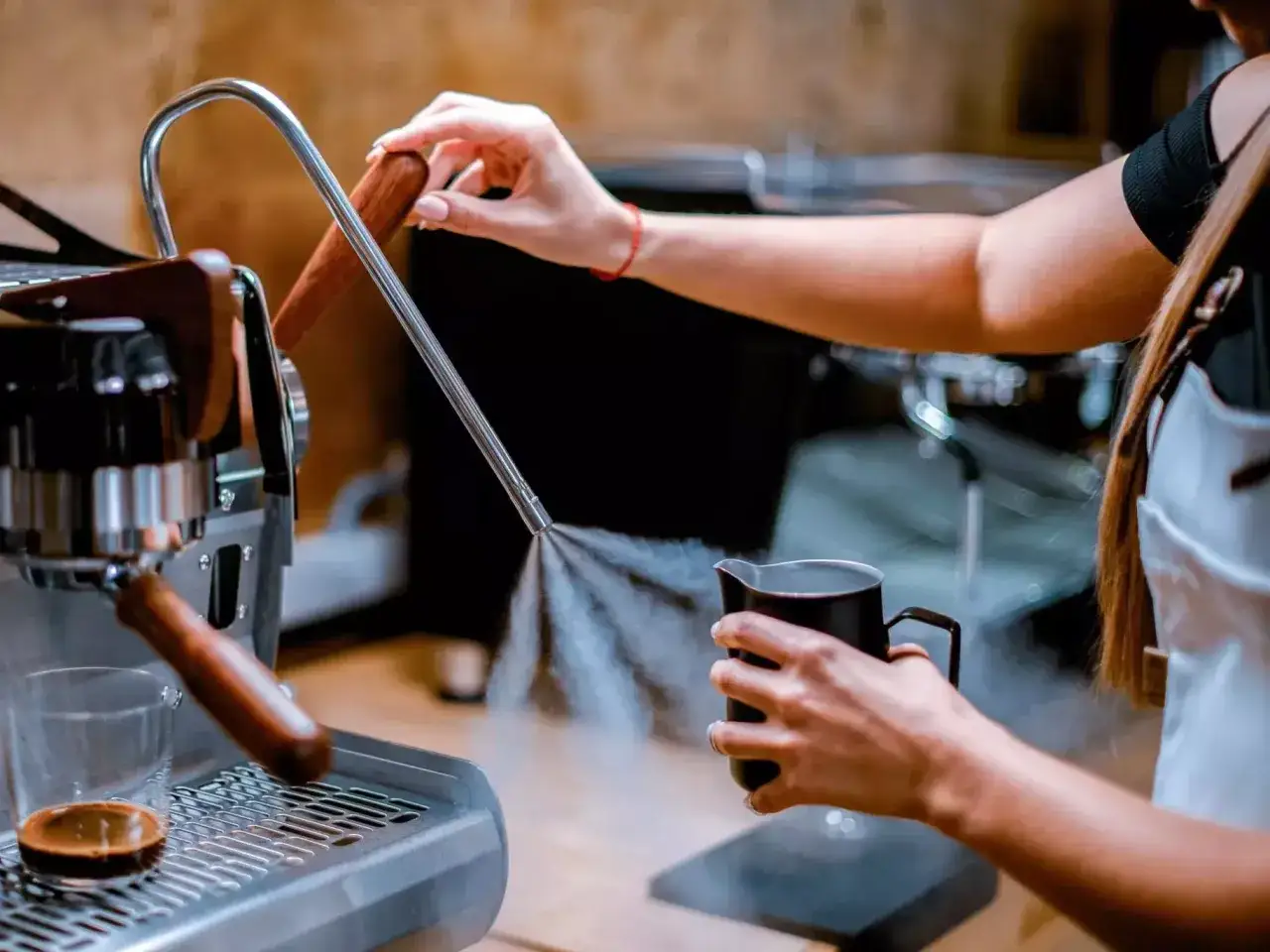 Barista reinigt die Espressomaschine mit Dampf. Der Dampfstrahl spr&uuml;ht aus der D&uuml;se in eine Kanne.