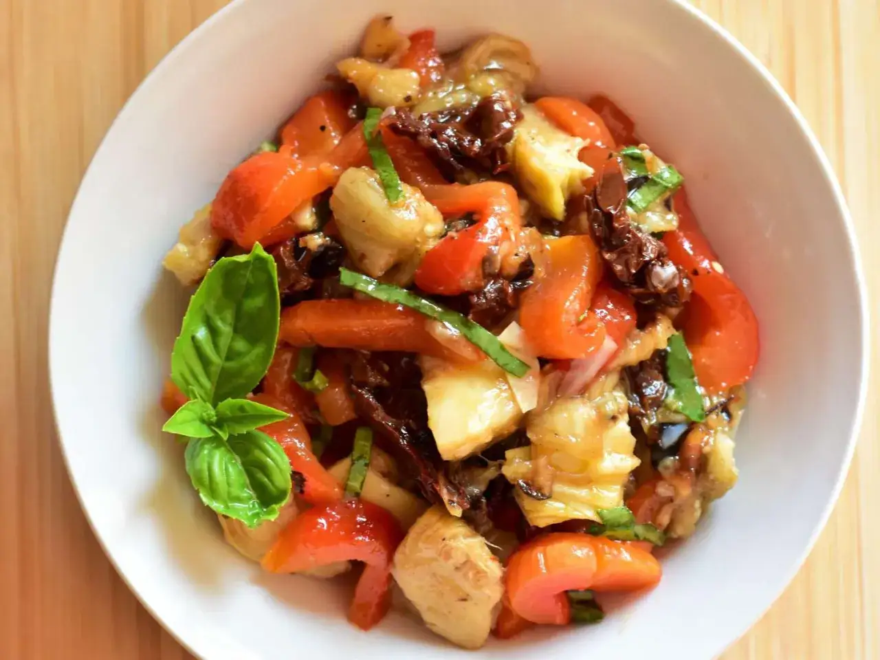 Ein köstliches Aubergine Paprika Rezept mit sonnengetrockneten Tomaten und frischem Basilikum in einer weißen Schüssel.