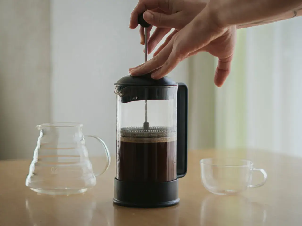 H&auml;nde dr&uuml;cken den Stempel einer French Press mit der perfekten Kaffeemenge herunter. Daneben stehen eine leere Glaskaraffe und eine Tasse.