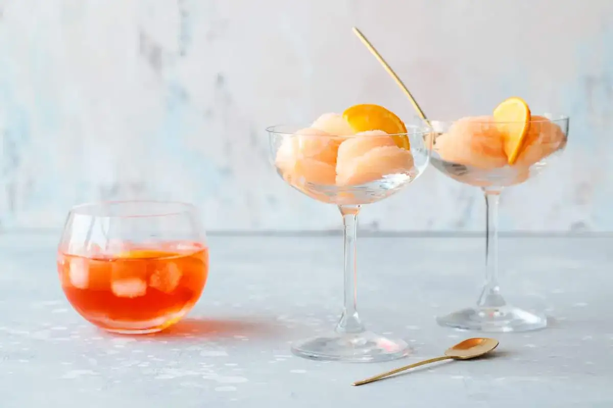 Erfrischendes Aperol Spritz Eis in eleganten Gläsern, garniert mit Orangenscheiben. Ein Aperol Spritz Cocktail steht daneben.