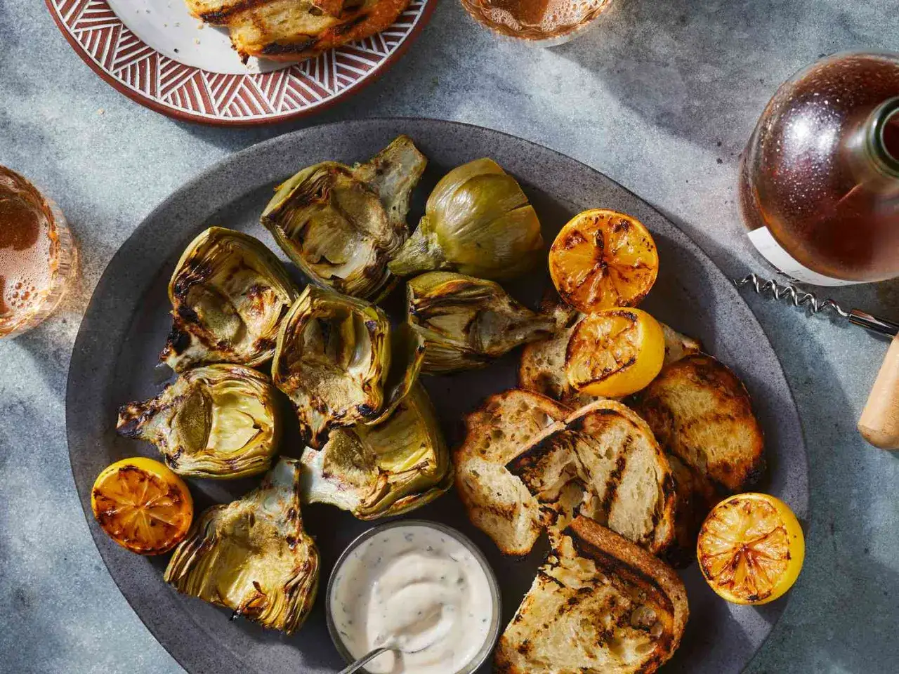 Artischocken grillen: K&ouml;stliche gegrillte Artischocken mit Zitronenspalten und Brot, serviert mit Dip.